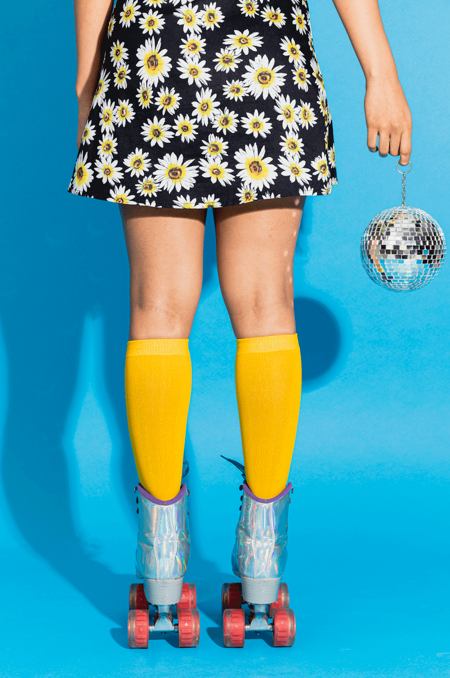 image of a woman standing against a blue background wearing rollerskates and holding a disco ball. She has a black skirt with white daisies on it and yellow knee high socks.