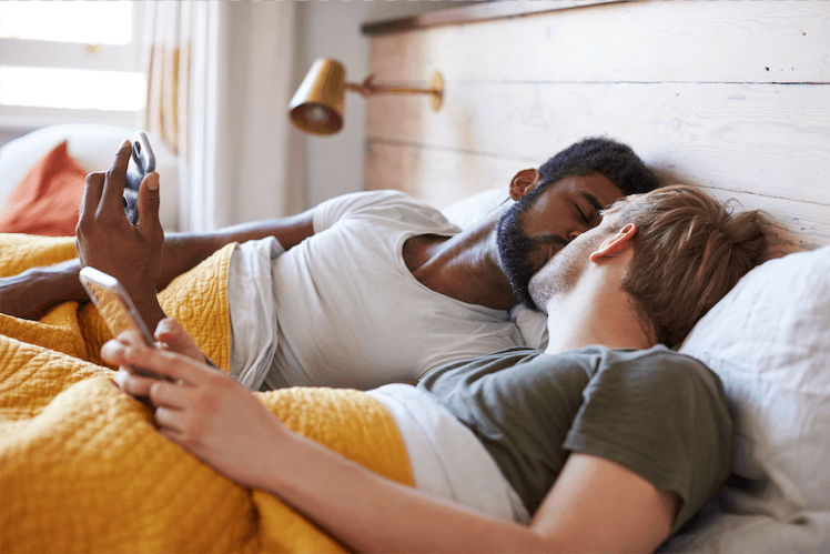 Two men (one Black, the other caucasian) lying in bed holding their phones and kissing.